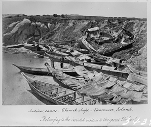 Pirogues de visiteurs venus participer à un potlatch dans l’Ancienne Réserve songhees en 1869. Trois pirogues de style salish de la Côte sont tirées sur le rivage à proximité d’une petite maison provisoire faite de nattes de joncs.