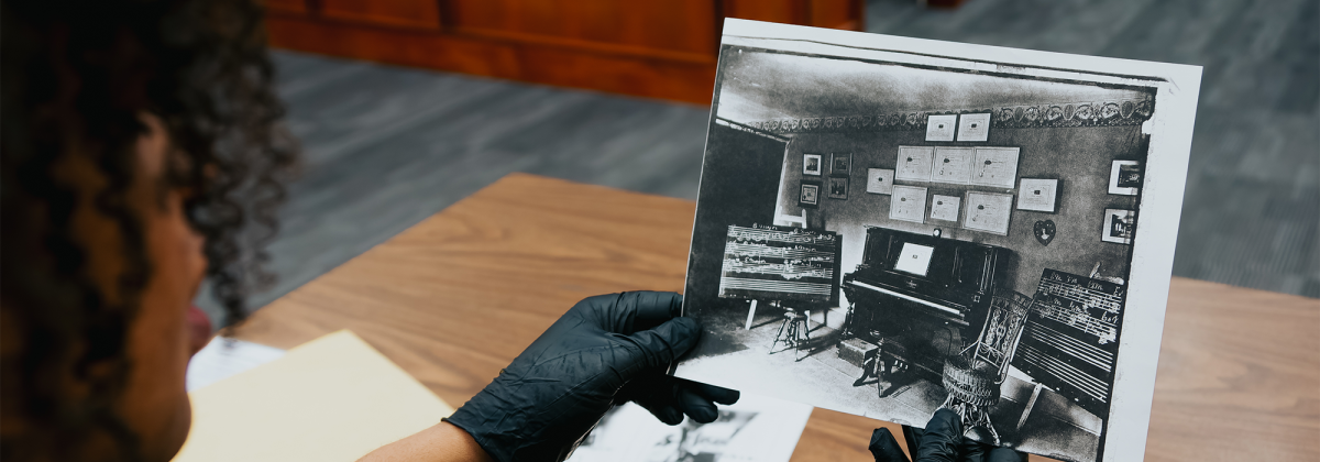 Gloved hands hold a black and white photograph of a music studio with a piano in the centre, and chalkboards with music notation on either side.